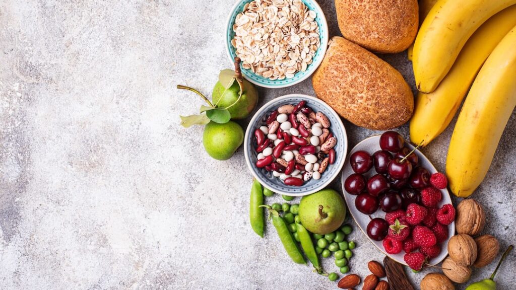 Healthy foods such as oatmeal, beans, fruit, bread and nuts on a stone surface.